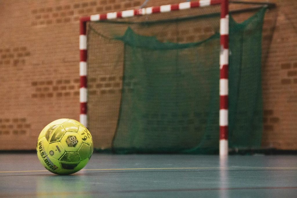 Bola amarela de futsal perto do gol com listras vermelhas em um ginásio esportivo.